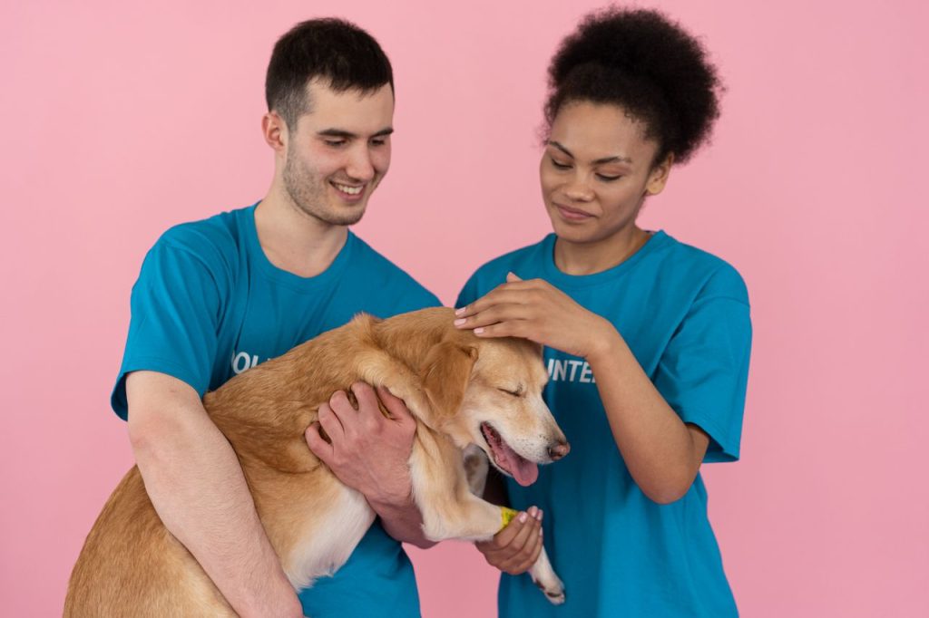 Two volunteers smile while petting a dog in a studio setting.